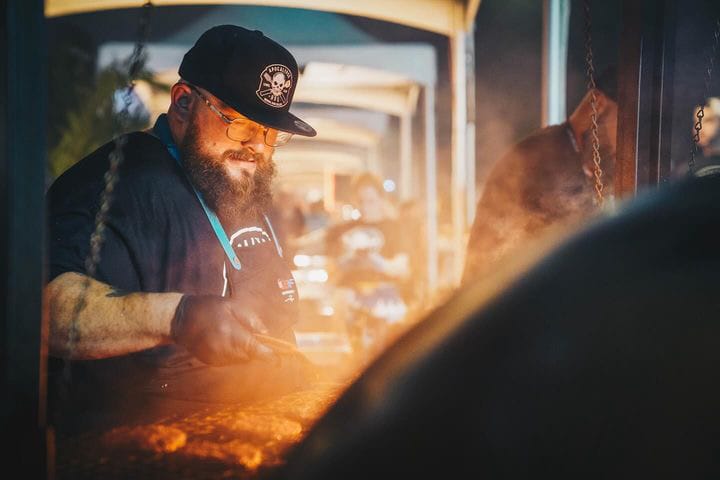 Jeff Budnechky, founder of Apocalypse BBQ, working the flat-top in an Apocalypse cap and apron as steam rises at dusk