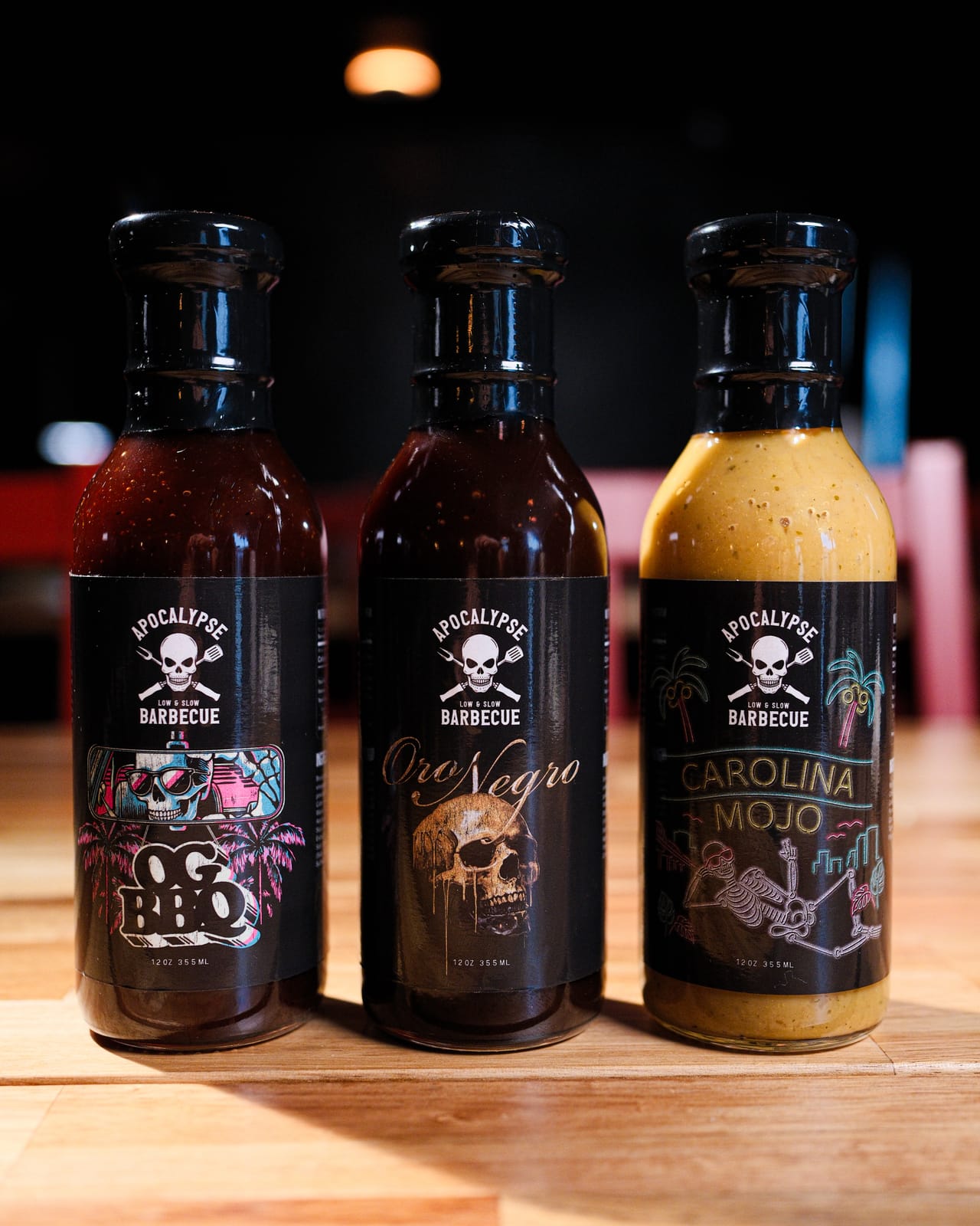 Three bottles of Apocalypse BBQ sauce — OG BBQ, Oro Negro, and Carolina Mojo — lined up on a wood table